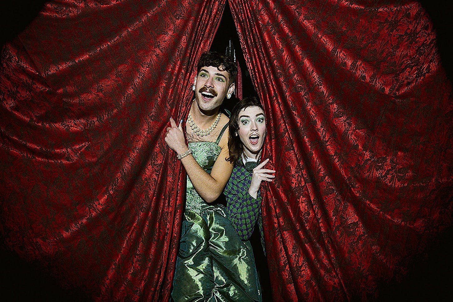 Two cabaret performers look out at the crowd from red velvet curtains.